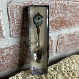 May include: A souvenir spoon in a box. The spoon has a decorative handle with a shield design and the words "Texas" and "Travel" on it. The spoon is silver-colored and the box is blue and white. The box reads "Made in USA".