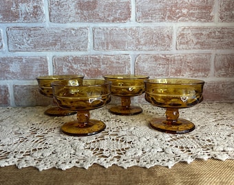 Può includere: Set di quattro coppette da dessert color ambra con base a piedistallo. I bicchieri hanno un motivo decorativo sul bordo e sono disposti su una tovaglia di pizzo davanti a un muro di mattoni.