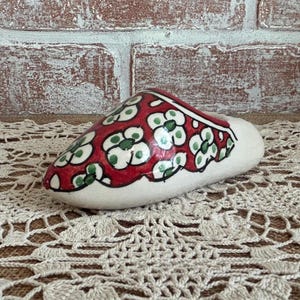 May include: A ceramic slipper-shaped decorative item. The slipper is white with a red top, adorned with green and white floral patterns. The item is placed on a beige lace doily, with a brick wall in the background.