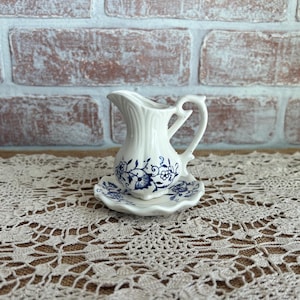 May include: A small white ceramic creamer with a handle and a matching saucer. The creamer and saucer are decorated with blue floral patterns. The creamer is sitting on a white lace tablecloth in front of a brick wall.