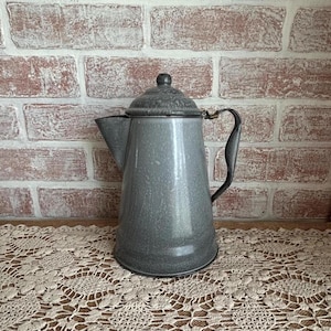 May include: Gray enamelware coffee pot with a lid and handle. The pot has a speckled finish and a spout for pouring. The background is a brick wall and a lace doily.