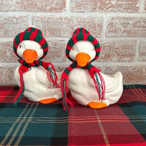 May include: Two white ceramic duck figurines wearing red and green striped knit hats and scarves. The ducks have orange beaks and feet. They are sitting on a red and green plaid tablecloth in front of a brick wall.