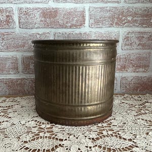 May include: A vintage, cylindrical metal container with vertical ribbed detailing. The container has a tarnished, aged appearance, with a dark rim and base. It sits on a cream-colored lace doily in front of a brick wall.
