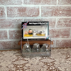 May include: A salt, pepper, and mustard set featuring three clear glass jars with silver-toned metal lids and a matching silver-toned metal wire stand. The set is displayed in front of its original cardboard box labeled Salt, Pepper & Mustard Set.
