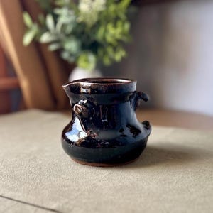 May include: A small, dark brown ceramic creamer with a glossy, reflective glaze. The vessel features a wide, rounded body, a distinct pouring spout, and two small, decorative loop handles on opposite sides. The base shows a raw, unglazed clay rim.