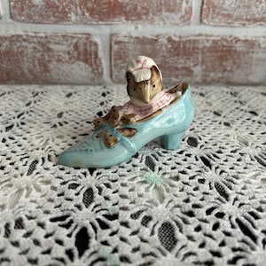 May include: A small ceramic figurine of a mouse wearing a pink dress and a white bonnet, sitting inside a blue shoe.
