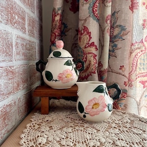 May include: A ceramic sugar bowl with a lid and a matching creamer pitcher, both featuring a cream-colored base with hand-painted pink flowers and dark green leaves. Each piece has a distinct dark green handle and a smooth, glossy finish.