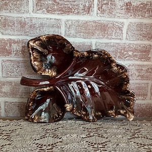 May include: A large, glossy, leaf-shaped ceramic dish in shades of brown and cream. The dish has a textured surface and a curved stem. The edges are ruffled, and the interior is smooth. The decorative piece is likely for display or serving.