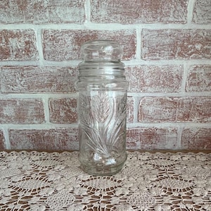 May include: A clear glass jar with a lid, featuring an embossed wheat stalk design. The jar is cylindrical and sits on a cream-colored lace doily. The background is a brick wall with a distressed, whitewashed finish.