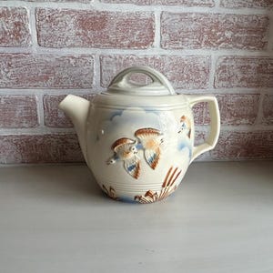 May include: Cream-colored ceramic teapot with a lid and handle. The teapot features a design of flying birds in shades of brown, blue, and white against a backdrop of clouds. The teapot is set against a brick wall.
