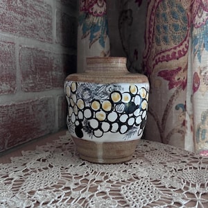 May include: A ceramic vase with a wide, rounded body and a short, narrow neck. The base and neck are unglazed tan clay, while the center features a white glaze decorated with a repeating pattern of black-outlined circles filled with yellow and black.