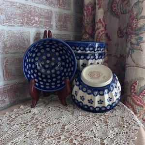 May include: Three ceramic bowls featuring a blue and white pattern. One bowl is displayed on a stand showing its interior with small blue circles, while two others are stacked, showing the exterior floral design and the Boleslawiec stamp.