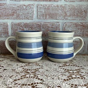 May include: Two ceramic mugs with a cream-colored base, blue and white horizontal stripes, and a blue interior. The mugs have a curved handle and are displayed on a lace tablecloth against a brick wall.