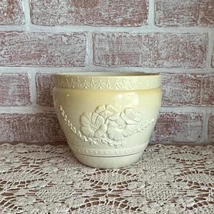 May include: Cream-colored ceramic planter with embossed floral design. The planter features a wide, rounded shape with a decorative band at the top and a floral motif on the front. The background is a brick wall.