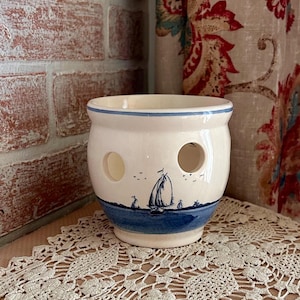 May include: A cream-colored ceramic yarn bowl featuring a blue painted nautical scene with a sailboat on the water. The bowl has a smooth, glazed finish with visible fine crackling and three circular cutouts along the upper side for yarn.