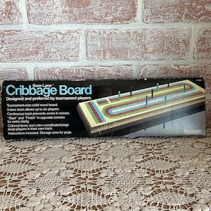 May include: A Sure-Lane Cribbage Board on a lace tablecloth. The board is tournament-sized with a 3-lane track for up to six players. The box text reads "Designed and preferred by tournament players."