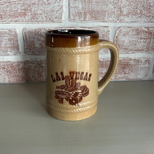 May include: Tan ceramic beer stein with a brown rim and handle. The stein features the words "LAS VEGAS" in a brown font, along with images of dice, poker chips, and a roulette wheel. The stein has a rope-like design around the top and bottom.