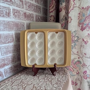 May include: A rectangular, tan-colored plastic serving tray featuring two white inserts, each containing eight oval-shaped indentations designed for holding eggs. The tray is supported by a dark brown, two-piece plastic display stand.