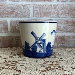 May include: A white ceramic planter with a blue windmill and landscape design. The planter has a dark blue rim and a detailed illustration of a windmill, trees, and clouds. The design is reminiscent of Delftware.