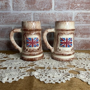 May include: Two vintage ceramic beer steins with handles. The steins are brown and cream-colored with a Union Jack flag on the front. The steins are sitting on a white lace doily.
