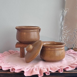 May include: Two brown speckled ceramic pots with a matching lid. One pot features a small pouring spout on the side. The pots have a rounded shape with a smooth finish and are positioned together, with the lid resting beside the second pot.