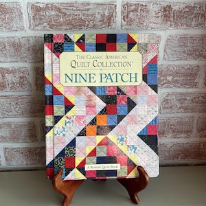 May include: A book titled "The Classic American Quilt Collection: Nine Patch" is displayed on a wooden stand. The book cover features a colorful patchwork design with red, blue, yellow, and green squares. The book is a Rodale Quilt Book.