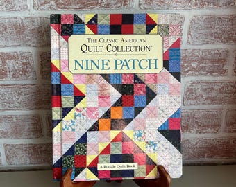 May include: A book titled "The Classic American Quilt Collection: Nine Patch" is displayed on a wooden stand. The book cover features a colorful patchwork design with red, blue, yellow, and green squares. The book is a Rodale Quilt Book.