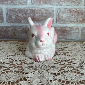 May include: A ceramic bunny figurine in white and pink. The bunny has pink ears, eyes, and accents. The figurine is sitting on a lace doily. The background is a brick wall.