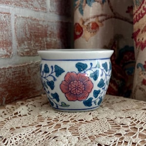 May include: A small, round, white ceramic bowl featuring a hand-painted floral pattern. The design includes a central pink flower with detailed petals surrounded by blue leafy vines. The bowl has a smooth, glossy finish and a flared rim at the top.