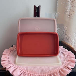 May include: A rectangular red plastic food storage container base positioned in front of two translucent, light pink rectangular lids. The items are stacked vertically, featuring smooth surfaces and rounded corners, with a circular indentation in the center of each lid and the base.