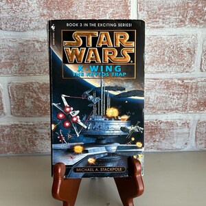 May include: A hardcover book titled "Star Wars X-Wing: The Krytos Trap" by Michael A. Stackpole. The cover features a space battle scene with X-wing fighters and a space station. The book is displayed on a brown wooden stand against a brick wall.