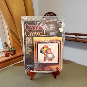 May include: A Columbia-Minerva Creative Crewel embroidery kit titled Lady Mouse 7310, featuring an Erica Wilson design. The package shows a finished crewel project of a mouse holding colorful balloons, enclosed in a clear plastic bag with text.