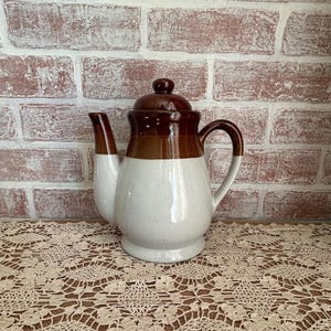 May include: A vintage ceramic teapot with a brown lid and handle, a brown band, and a speckled white body. The teapot has a curved spout and sits on a lace doily in front of a brick wall.