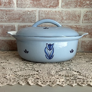 May include: A light blue enamel Dutch oven with a matching lid and handle. The pot features a blue tulip design on the front and small floral accents. The pot is sitting on a beige lace tablecloth.