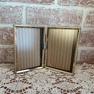 May include: A vintage gold-toned metal double picture frame. The frame is open, revealing the cardboard backing. The frame is rectangular with a decorative edge. The background is a brick wall.