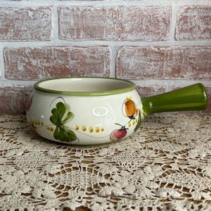May include: A white ceramic soup bowl with a green handle and rim. The bowl is decorated with painted green leaves, orange and red fruit, and small yellow dots. The bowl is sitting on a lace tablecloth.
