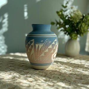 May include: A ceramic vase featuring a gradient color scheme that transitions from blue at the top to a light tan at the base. The surface is decorated with white etched geometric patterns and horizontal lines encircling the neck and bottom.