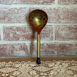 May include: A decorative wooden spoon with a red bowl and a gold handle. The bowl features a gold floral design. The spoon is set against a brick wall and a lace tablecloth.