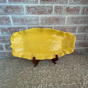 May include: A bright yellow, leaf-shaped ceramic serving platter. The platter is supported by a brown wooden stand. The background features a brick wall and a lace tablecloth.