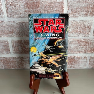 May include: A vintage paperback book titled "Star Wars X-Wing Rogue Squadron" by Michael A. Stackpole. The cover features a space battle scene with X-wing fighters and the Star Wars logo in red and white against a blue background. The book is displayed on a wooden stand.
