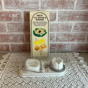 May include: A white plastic multi egg slicer and wedger with a clear plastic lid. The product is displayed in front of a brick wall. The product's box is visible, with the text "MULTI EGG SLICER & WEDGER".