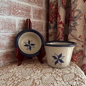 May include: A cream-colored ceramic bowl and a matching ceramic mug, both featuring a dark blue eight-pointed star pattern. The bowl is displayed on a small wooden stand, while the mug sits beside it, showcasing a smooth, glazed finish.