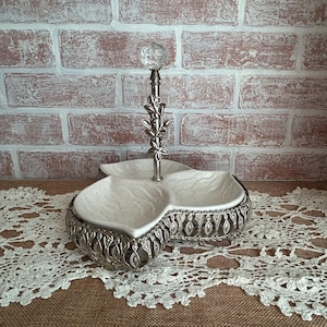 May include: A three-tiered serving dish with a silver-colored metal frame and a clear glass ball on top. The dish has three sections with a speckled white interior and a decorative floral design.