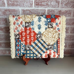 May include: A quilted patchwork pillow cover with a cream-colored ruffle trim. The cover features a mix of floral and geometric patterns in red, blue, beige, and black on a white background. The pillow is displayed on a wooden stand.