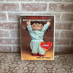 May include: A vintage Cuddle Kids soft sculpture doll kit. The box features a doll in a light blue outfit with red shoes, arms raised. The text reads "Cuddle Kids Soft Sculpture Doll Kit" and "Full 20" Doll".