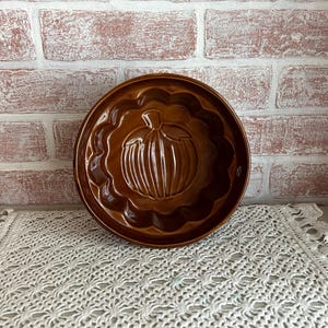 May include: A brown ceramic pumpkin mold with a scalloped edge. The pumpkin design is embossed in the center of the mold. The mold is sitting on a white lace doily in front of a brick wall.