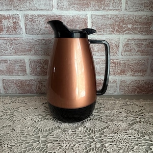May include: A copper-colored insulated pitcher with a black handle, lid, and base. The pitcher has a sleek, rounded shape and is designed for serving beverages. The background is a brick wall.