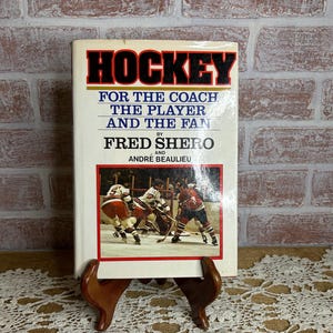 May include: A vintage book titled "HOCKEY" with the subtitle "FOR THE COACH, THE PLAYER AND THE FAN." The cover features a hockey game image and the authors' names, Fred Shero and André Beaulieu. The book is displayed on a wooden stand.