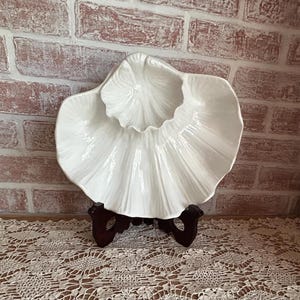 May include: A white, shell-shaped ceramic serving dish with a ruffled edge, displayed on a dark wooden stand. The dish has a textured, ribbed design and sits on a lace tablecloth against a brick wall.