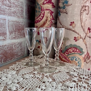 May include: Three clear glass cordial glasses with slender bowls and stems featuring a series of stacked spherical beads. Each glass has a circular base and a smooth, transparent finish, standing upright in a row on a flat surface.